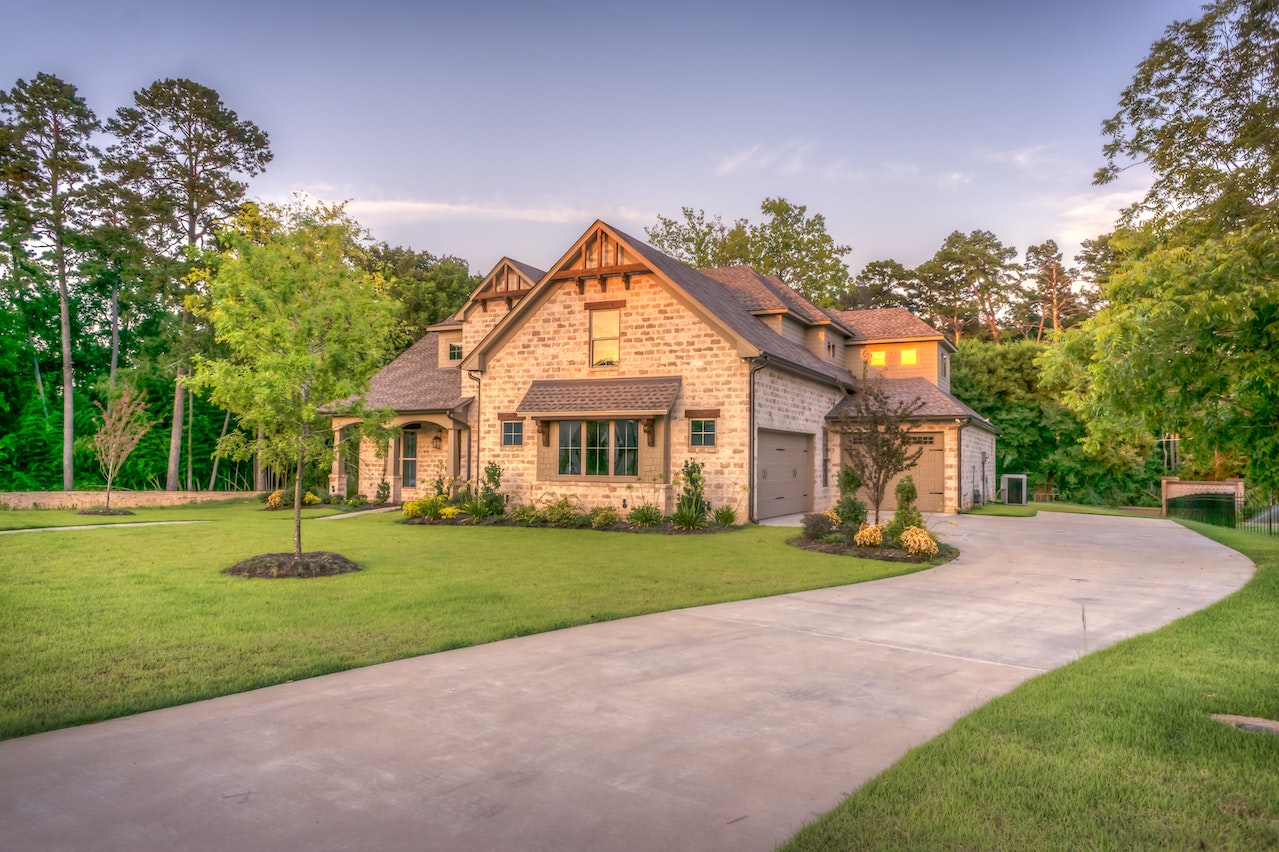 A large stone house with a manicured lawn is surrounded by trees. The property, ideal for real estate investment companies, features multiple gables, large windows, and green shutters, with a paved curved driveway leading to an attached garage. The sky is clear and the scene is well-lit by natural light. | MONEY6X