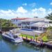 A luxurious waterfront property featuring a large modern house with a white exterior, expansive windows, and a spacious outdoor pool. This best real estate investment offers a private dock with boats moored, set against lush greenery and a serene waterway under a bright blue sky. | MONEY6X