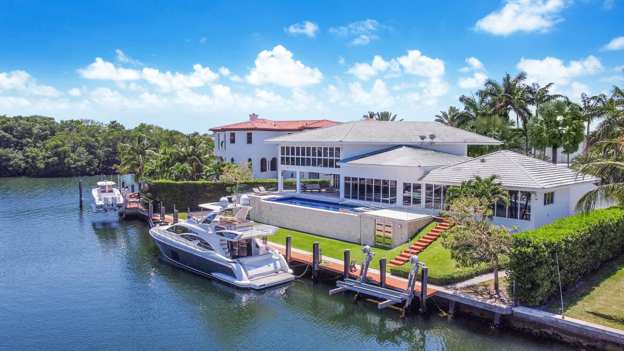 A luxurious waterfront property featuring a large modern house with a white exterior, expansive windows, and a spacious outdoor pool. This best real estate investment offers a private dock with boats moored, set against lush greenery and a serene waterway under a bright blue sky. | MONEY6X