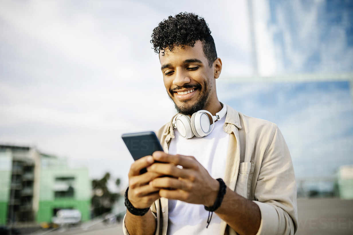 A smiling person with curly hair and wearing headphones around their neck is holding and looking at a smartphone, possibly exploring apps to make money. They are dressed in a beige jacket over a white shirt, standing outdoors with a blurred background of buildings and a blue sky. | MONEY6X