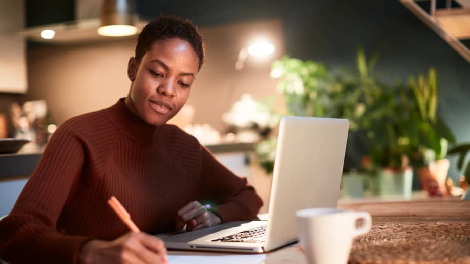 A person with short hair is sitting at a table in a cozy, dimly-lit room, using a laptop and writing notes on paper. They are wearing a brown sweater, and a coffee mug is placed on the table beside them. There are blurred plants in the background as they focus on applying the 50/30/20 Rule. | MONEY6X