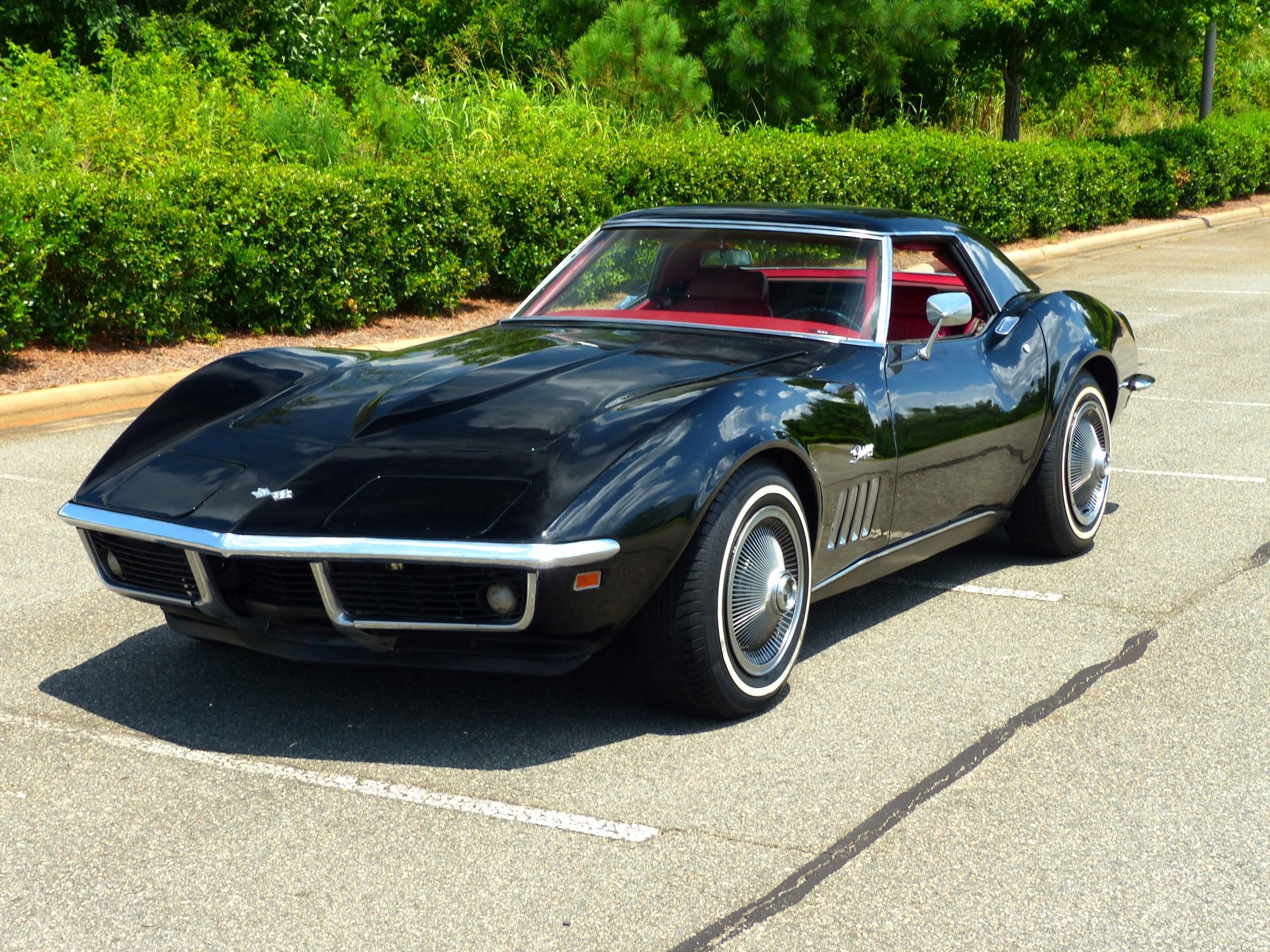 A sleek, black classic car with a low profile and stylish curves is parked in a lot. Its polished body shines under the sunlight, highlighting its vintage design. The car has a red interior, and a wooded area with greenery can be seen in the background—a true gem among classic cars. | MONEY6X