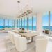 A modern dining area in an upscale apartment features a wooden dining table surrounded by white chairs. A geometric chandelier hangs above. Floor-to-ceiling windows, virtually staged by top companies, offer a stunning ocean view with clear blue skies and sunlight illuminating the room. | MONEY6X