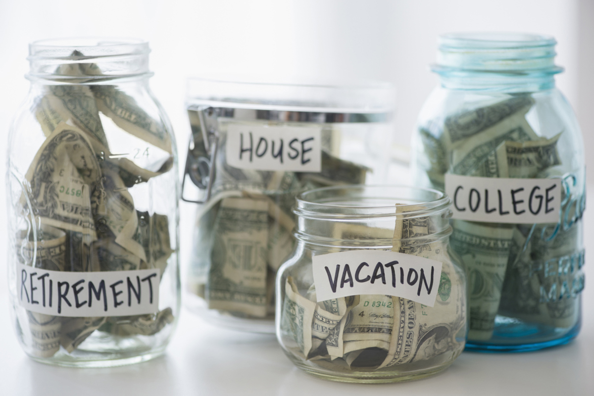Four glass jars filled with U.S. dollar bills are labeled “Retirement,” “House,” “Vacation,” and “College.” The labels, written on pieces of paper taped to the jars, signify savings goals. These jars, arranged in a line against a light background, illustrate some of the best ways to save money effectively. | MONEY6X