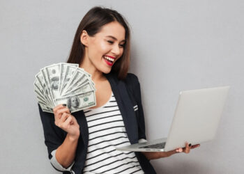 A happy woman wearing a blazer and striped shirt is holding multiple U.S. hundred-dollar bills in one hand while using a laptop with the other. She is smiling and standing against a gray background, showcasing how to make money online. | MONEY6X