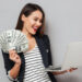 A happy woman wearing a blazer and striped shirt is holding multiple U.S. hundred-dollar bills in one hand while using a laptop with the other. She is smiling and standing against a gray background, showcasing how to make money online. | MONEY6X