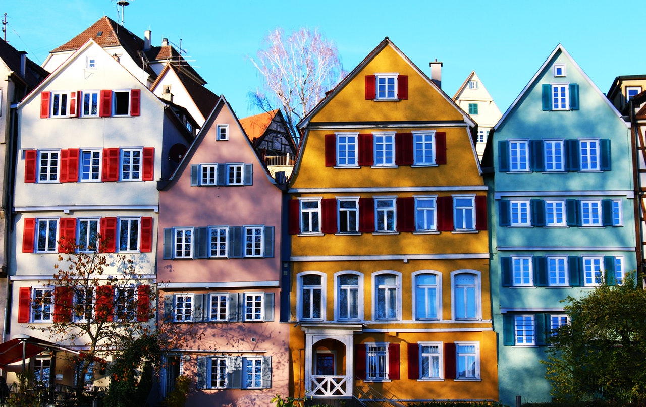 A row of colorful, traditional European houses stands under a clear blue sky. The buildings, perfect for Germany for real estate investment, are painted in shades of blue, pink, yellow, and green with contrasting window shutters. Trees and shrubs can be seen in the foreground. | MONEY6X