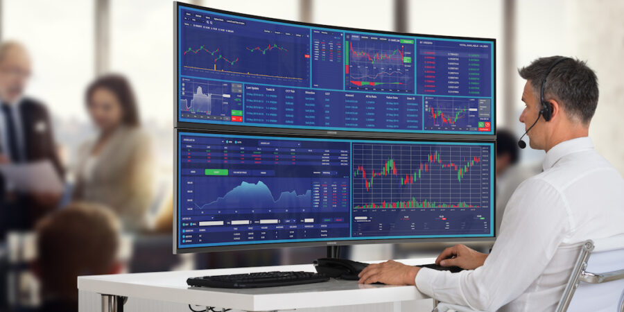 A man wearing a headset is seated at a desk in front of two large computer monitors displaying various financial charts and market monitoring tools. In the blurred background, two people are having a conversation. | MONEY6X