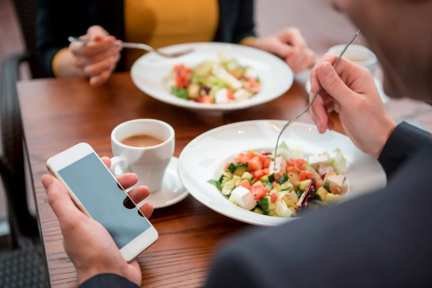 Two individuals are seated at a wooden table, enjoying fresh salads and cups of coffee. One person is holding a fork in one hand and a smartphone in the other, perhaps checking food expenses, while the other is just about to take a bite. The scene suggests a casual, modern dining experience. | MONEY6X