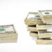 A photo showcases multiple stacks of U.S. hundred-dollar bills. On the left, there is a smaller stack lying flat, while the right side features several larger money stacks, bound with white paper bands, arranged vertically. | MONEY6X
