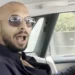 A person with a shaved head and sunglasses is sitting in a car, possibly pondering How Did Andrew Tate Make His Money. They are wearing a dark jacket and have their mouth open as if speaking. The car interior appears luxury with a beige headrest, and there is a cityscape visible through the window. | MONEY6X