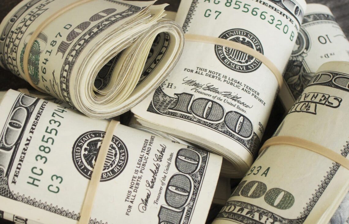 Several bundles of rolled-up U.S. 100-dollar bills secured with rubber bands are piled together, equating to money 6x over. Each bundle includes multiple 100-dollar bills and displays the recognizable face and design of American currency. | MONEY6X