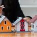 A person holding a pen points toward three miniature house models on a wooden desk. The models, in yellow, red, and white with distinct architectural designs, represent various real estate options. The person is partially visible, focusing intently on the houses. | MONEY6X