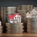 Three miniature houses sit atop stacks of coins of varying heights. The houses are detailed, with the first featuring a blue roof, the second with a red roof, and the third with a beige roof. The image symbolizes the concept of real estate investment and money in 6x REIT holdings. | MONEY6X