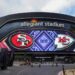A wide view of Allegiant Stadium with a large electronic display featuring the logos of the San Francisco 49ers and the Kansas City Chiefs, indicating an upcoming game between the two teams. The stadium exterior is sleek and modern with visible walkways and plants—imagine this excitement when you attend Super Bowl 2024. | MONEY6X
