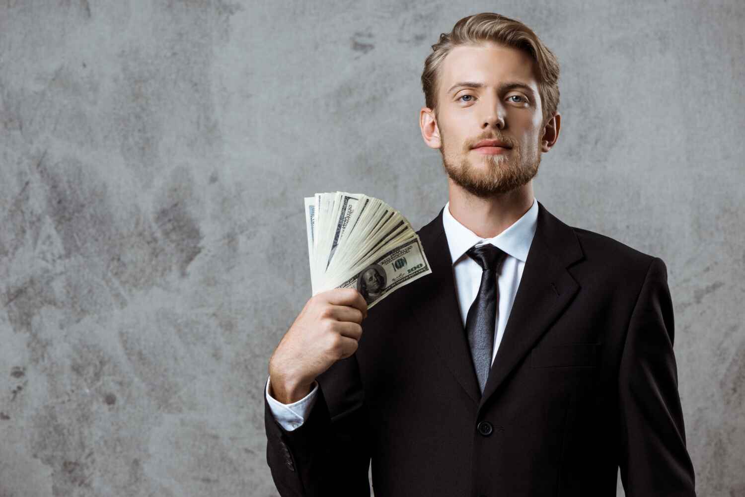 A man wearing a black suit and tie stands against a gray, textured background. He holds several fan-spread U.S. dollar bills in one hand near his face, looking directly at the camera with a neutral expression, as if contemplating ways to invest money. | MONEY6X