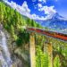 A bright orange train travels across a tall, narrow bridge surrounded by lush greenery and pine trees. A cascading waterfall flows down the cliffside on the left. Snow-capped mountains rise in the background under a bright, cloud-filled blue sky, capturing the essence of Luxury Swiss Vacations. | MONEY6X