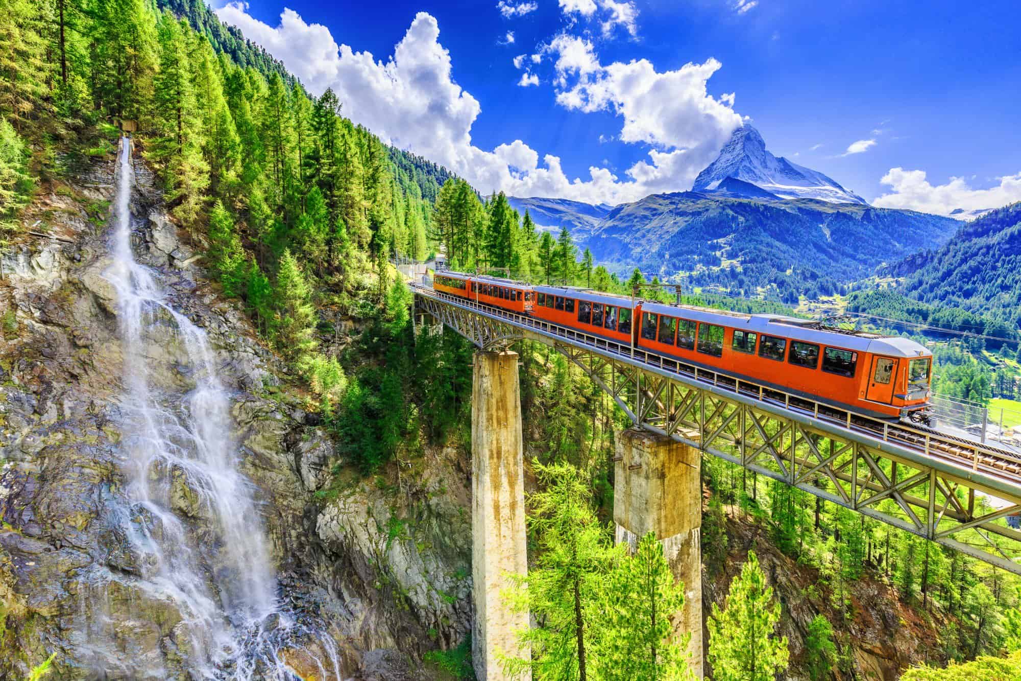 A bright orange train travels across a tall, narrow bridge surrounded by lush greenery and pine trees. A cascading waterfall flows down the cliffside on the left. Snow-capped mountains rise in the background under a bright, cloud-filled blue sky, capturing the essence of Luxury Swiss Vacations. | MONEY6X