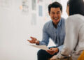 A man in a blue shirt is sitting and smiling while holding a paper, engaging in a conversation with a woman with long dark hair who is facing away from the camera. They are in a well-lit room with blurred documents and notes on the wall, possibly discussing hiring details. | MONEY6X