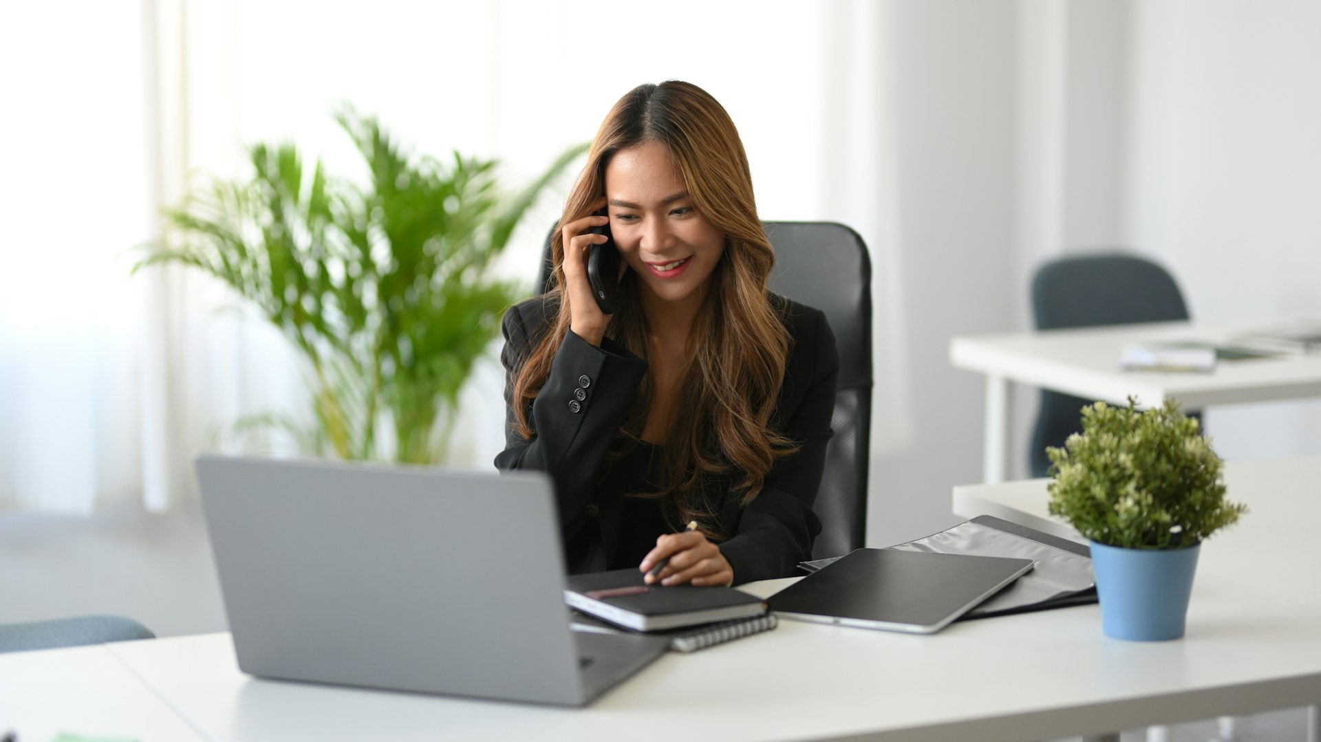 A woman with long hair sits at a desk in a modern office, smiling while talking on the phone. She is surrounded by a laptop, notebooks, and a potted plant as she discusses earnings online. A large leafy plant near a bright window adds to the vibrant atmosphere. | MONEY6X