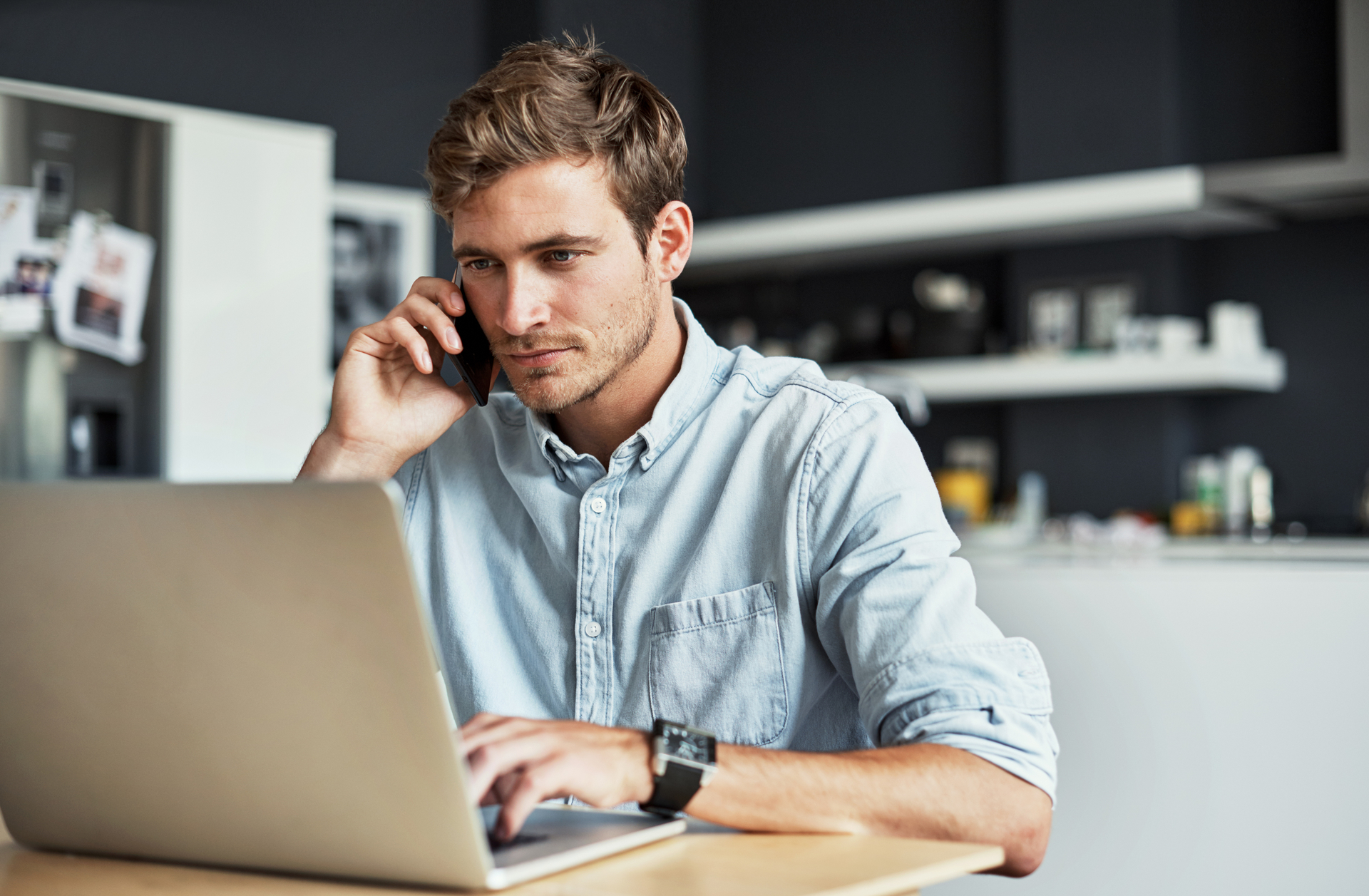 A man with short, light brown hair and a beard is sitting at a table, looking at a laptop screen while talking on a smartphone about invest opportunities. He is wearing a light blue button-up shirt and a wristwatch. Behind him, a modern kitchen is visible in the blurred background. | MONEY6X