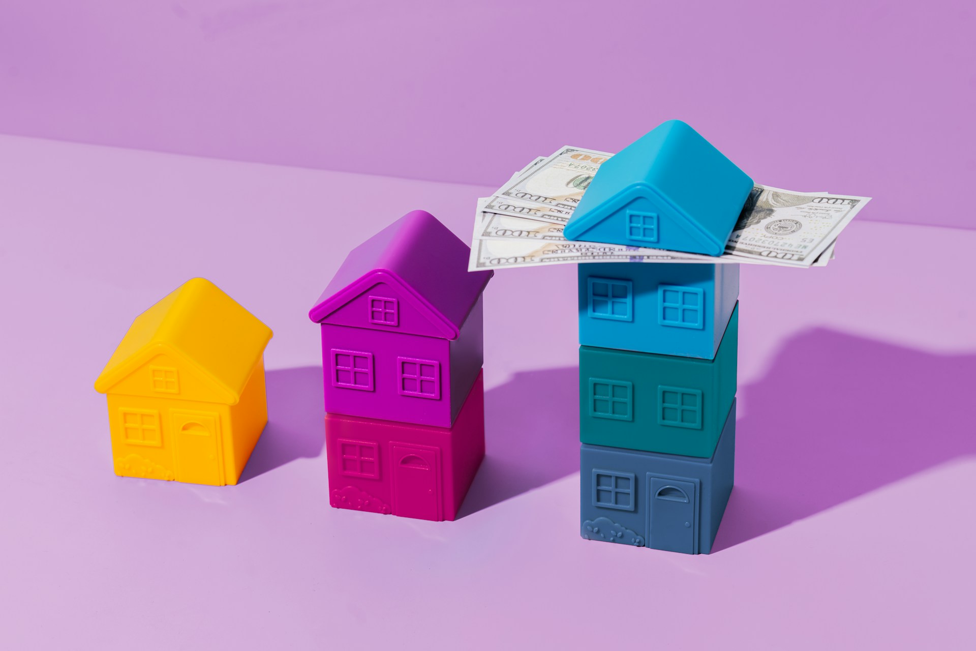 Three colored toy houses are stacked progressively taller from left to right (yellow, purple, and multiple shades of blue). The tallest stack of blue houses has several U.S. dollar bills resting on the roof against a purple backdrop, subtly hinting at Money6x.com. | MONEY6X