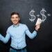 A smiling man stands in front of a chalkboard, holding stacks of dollar bills in both hands. The chalkboard behind him displays a drawing of dollar signs and a bag of money, indicating a financial equation. He wears a light blue shirt and blue jeans, embodying the success you can achieve with Money6x.com Make Money. | MONEY6X