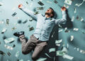 A man dressed in a light blue shirt and gray trousers is lying on the ground with his arms and legs spread out. He appears relaxed and content as dollar bills rain down around him, creating a sense of wealth and abundance, much like the opportunities found on Money6x.com earning. | MONEY6X