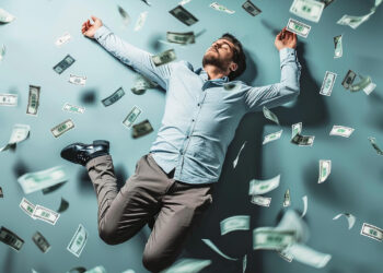 A man dressed in a light blue shirt and gray trousers is lying on the ground with his arms and legs spread out. He appears relaxed and content as dollar bills rain down around him, creating a sense of wealth and abundance, much like the opportunities found on Money6x.com earning. | MONEY6X