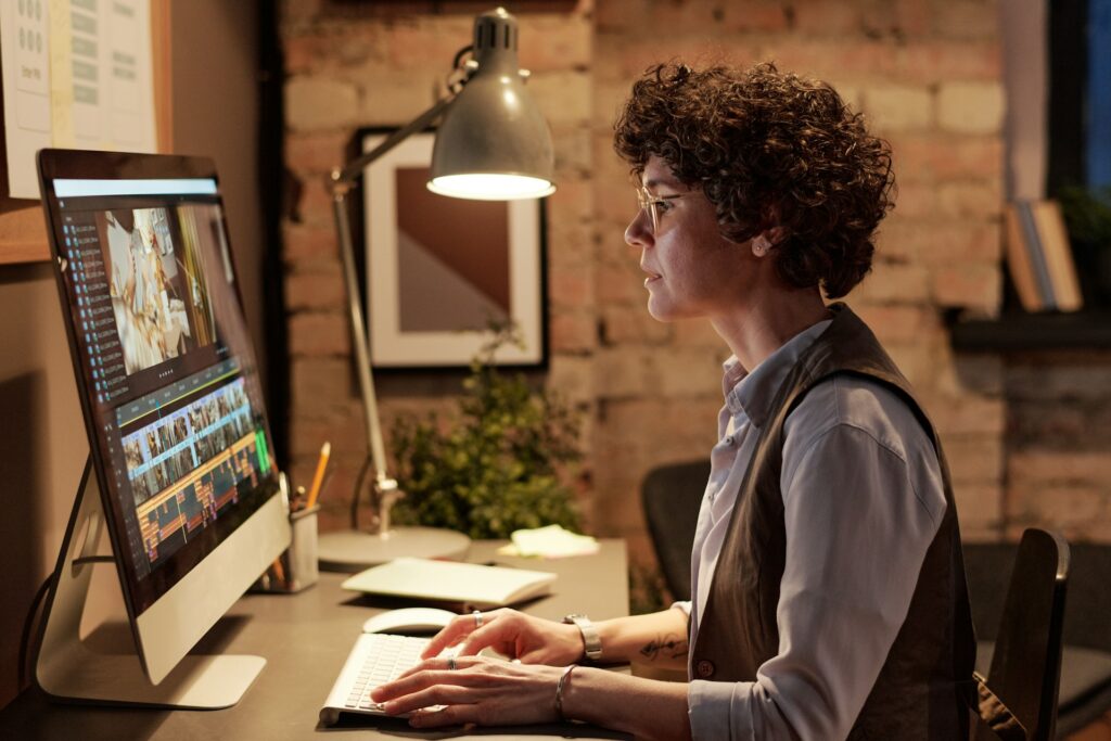 A person with curly hair and glasses is seated at a desk, editing a video on a large computer monitor. The background features exposed brick walls and framed pictures. Desk items include a lamp, pencils, a plant, and a keyboard bearing the logo of Money6x.com. The setting appears cozy and creative. | MONEY6X