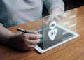 A person is holding a stylus pen, interacting with a transparent digital interface on a tablet. The interface displays an AI icon and speech bubbles, suggesting a chat with artificial intelligence to improve your financial literacy. | MONEY6X