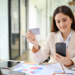 A woman in a beige blazer sits at a desk with a smartphone in one hand and a credit card in the other. Amongst the laptop, tablet, and colorful charts, she appears happy—perhaps exploring core banking solutions while conducting an online transaction. | MONEY6X