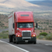 A red semi-truck, highlighting the journey of an owner-operator managing expenses, cruises along a highway in a desert landscape. The road curves with hills and sparse vegetation visible under a partly cloudy sky. Another truck is partially captured on the right side of the image. | MONEY6X