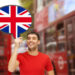 A man in a red shirt holds a speech bubble adorned with the British flag, standing amidst blurred red double-decker buses on a bustling street, contemplating how to start a business in the UK. | MONEY6X
