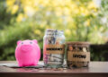 A pink piggy bank, a jar labeled "Emergency Fund" filled with dollar bills, and a jar labeled "Saving" brimming with coins sit on a wooden table. Several coins and U.S. dollar bills are scattered nearby, while blurred greenery provides the backdrop. | MONEY6X