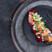 A black plate with smoked salmon, beet slices, cucumber, figs, and crumbled feta topped with microgreens. The dish is artistically arranged on a metal tray with handles, placed on a dark textured surface. | MONEY6X