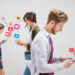 Three people walk while looking at their phones, surrounded by floating social media icons and notifications like hearts, likes, and emojis. This scene illustrates how using social media for your business can boost digital engagement and interaction. | MONEY6X