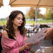 A woman in a pink jacket sits at an outdoor café table, reading from menu holders. A person hands the menu to her. Glasses of water are on the table, and patio umbrellas and greenery provide a serene background. | MONEY6X