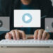Person typing on a white keyboard, three floating video play icons above the keys symbolizing online video content creation or streaming with a focus on refined video content strategy. Dressed in a black blazer, the setting is a straightforward office desk setup. | MONEY6X