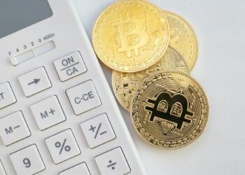 A white calculator is positioned next to three gold-colored coins with the Bitcoin symbol, hinting at the complexities of crypto taxation. The coins boast an intricate circuit-like design, emphasizing a focus on cryptocurrency and finance against a plain, minimalistic background. | MONEY6X