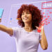 A woman with curly red hair takes a selfie with her smartphone, aiming to go viral on social media. Virtual icons like "LIVE," hearts, and comments float in front of her against a vibrant purple background. | MONEY6X