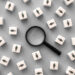 A magnifying glass sits among white cubes with minimalist human icons, some upside down, on a gray background. The image conveys a sense of searching to find someone or conducting thorough analysis. | MONEY6X
