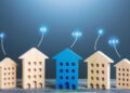 Five wooden house models stand in a row on a table, with the central house painted blue. Euro symbols (€) float above each house, representing housing prices or real estate value—ideal for illustrating passive income opportunities. Blue gradient background. | MONEY6X