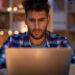 A young man with a beard looks intently at a laptop screen in a cozy, dimly lit room decorated with string lights—capturing the quiet focus of people trying to make money online. Shelves line the background. | MONEY6X