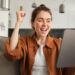 A young woman sitting on a couch in a modern living room smiles and raises her fist in excitement while looking at a tablet, thrilled to discover new ways to earn money from home. | MONEY6X