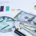 A magnifying glass, spreadsheets, colorful charts, and several U.S. $100 bills on a desk, representing financial analysis, business reports, money management, and personal wealth creation. | MONEY6X