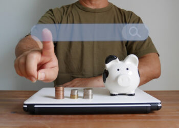 A person in a brown shirt touches a virtual search bar above a laptop, with a piggy bank and stacks of coins on the laptop, symbolizing online financial searches or exploring Top Alternatives to Traditional Personal Loans. | MONEY6X