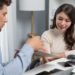A man hands a document to a smiling woman sitting at a desk with a calculator, smartphone, and papers. They appear to be discussing work or finances, possibly using a broker to help get the best deals on loans in a modern office setting. | MONEY6X