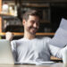 A man sitting at a table with a laptop and coffee cup smiles and raises his fist in excitement while reading a document, showing the satisfaction of getting more from every invoice you send as a freelancer. | MONEY6X