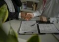 Two people in formal attire shake hands across a desk with documents, a clipboard, and a pen, suggesting an Installment Loans agreement or contract signing. A green plant is visible in the foreground. | MONEY6X